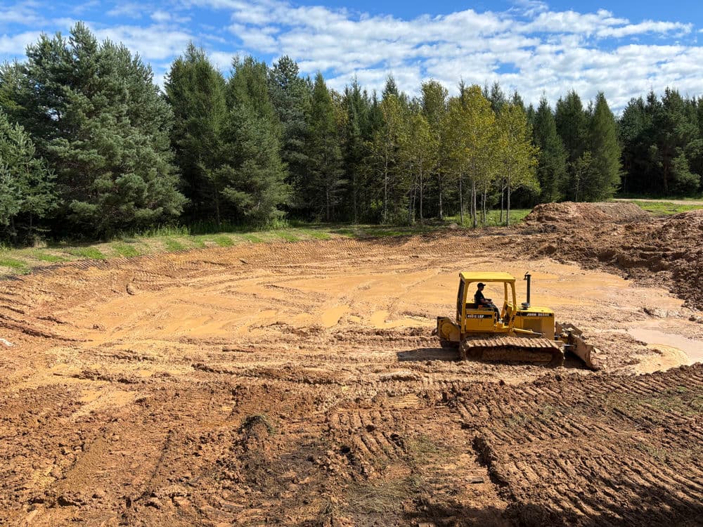 Bulldozer on construction site with trees, digging and leveling soil under a blue sky.