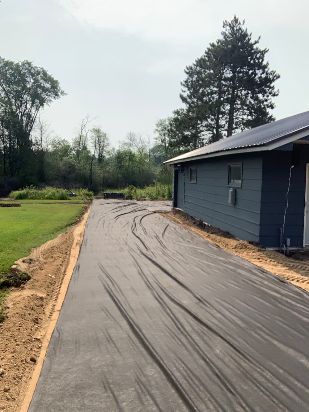 Freshly paved black asphalt driveway leading to a modern blue house in a rural setting.